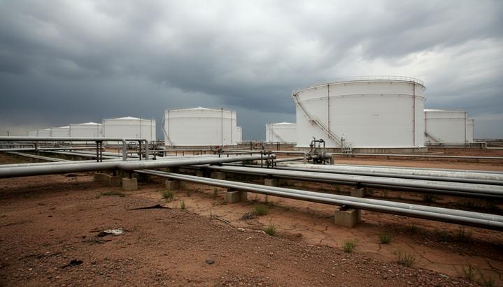 Крупные белые резервуары для хранения нефти на нефтебазе, соединенные сетью трубопроводов, на фоне пасмурного неба.