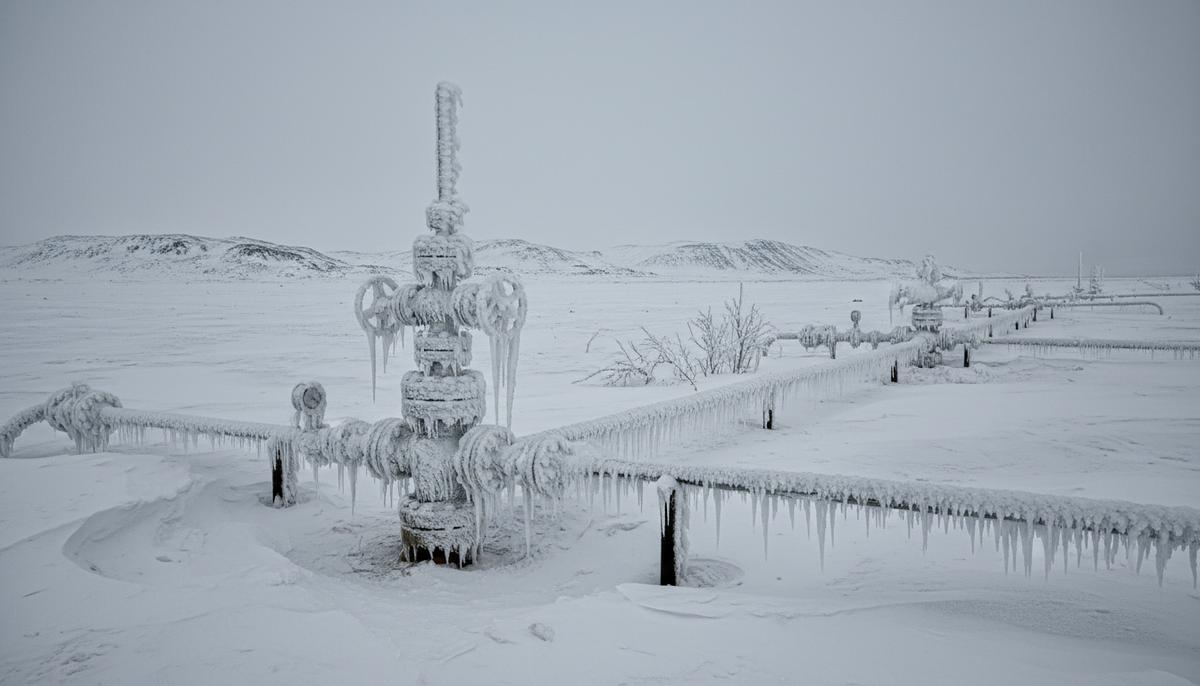 Газовый трубопровод и оборудование скважины, покрытые толстым слоем инея и льда на фоне заснеженного поля в пасмурную погоду.